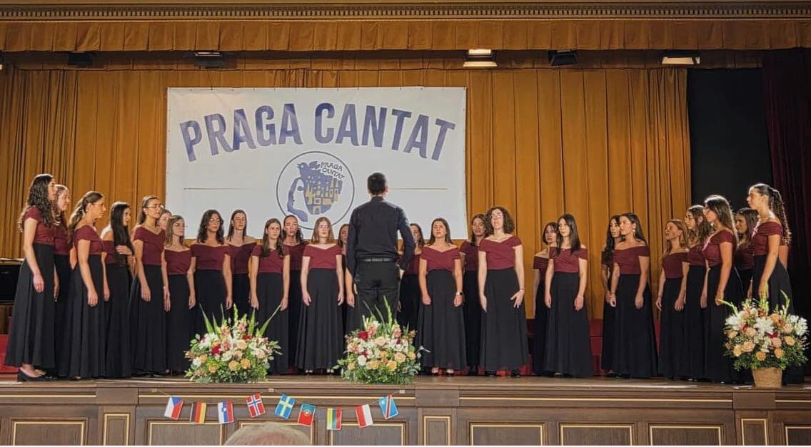 Coro feminino em palco, debaixo de uma faixa com o nome do festival Praga Cantat
